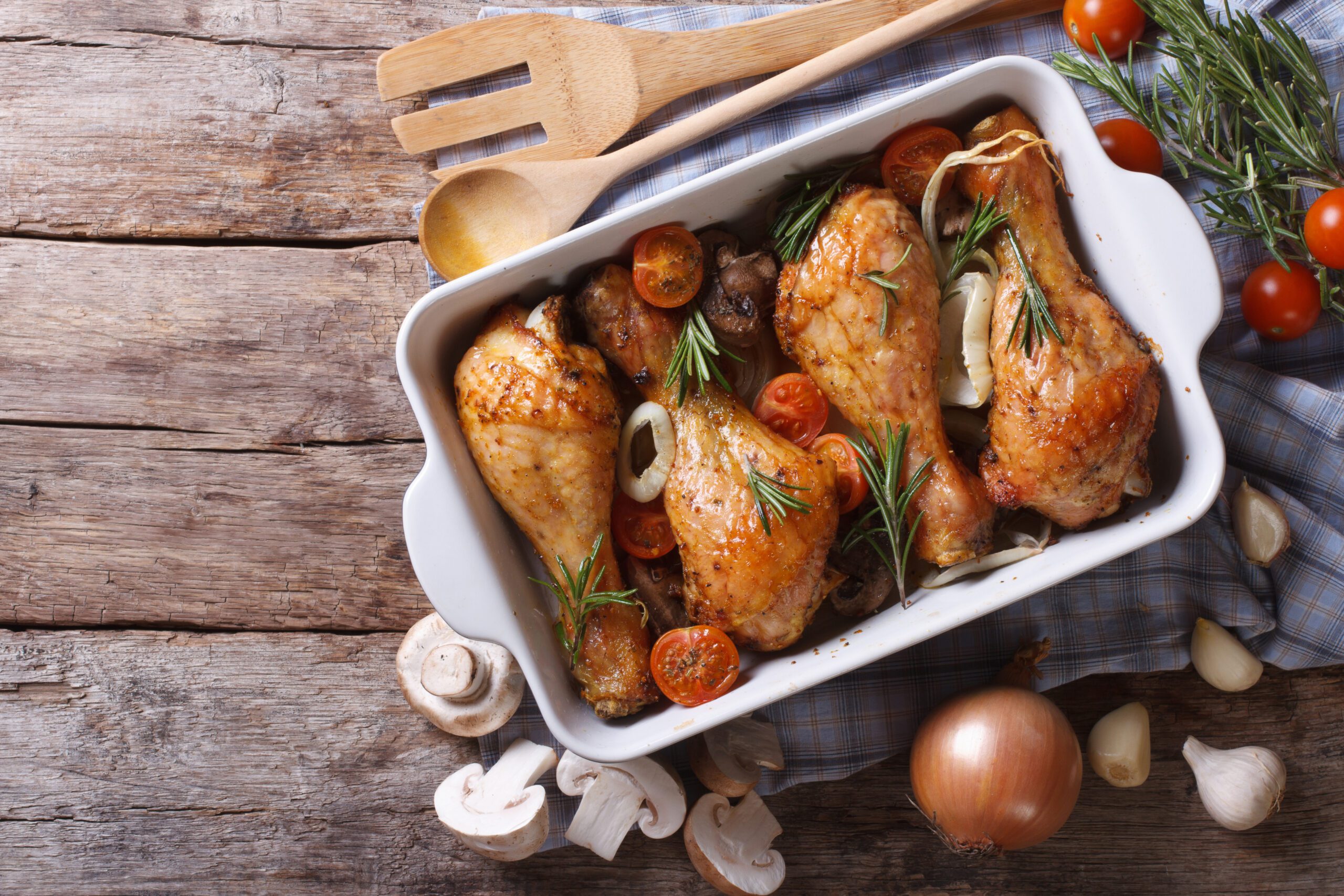 Baked chicken drumsticks with cherry tomatoes, mushrooms, onions, and rosemary in a white casserole dish on a wooden table with cooking utensils.