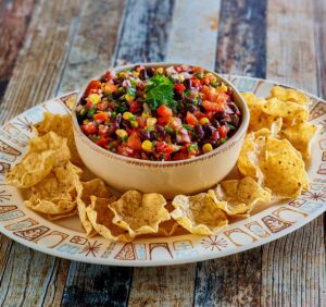 A bowl of salsa with black beans, corn, tomatoes, and herbs, surrounded by tortilla chips on a decorative plate.