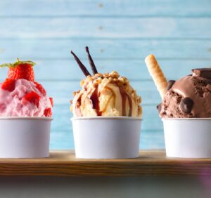 Three cups of ice cream on a wooden surface: strawberry with strawberries, vanilla with nuts and syrup, and chocolate with a wafer stick. Blue wooden background.