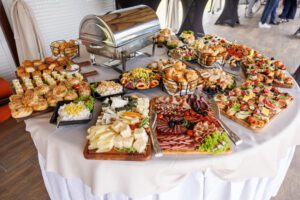 A round table covered with a white cloth displays a buffet of appetizers, including cheese, meat, sandwiches, and salads, with a metal chafing dish at the center.