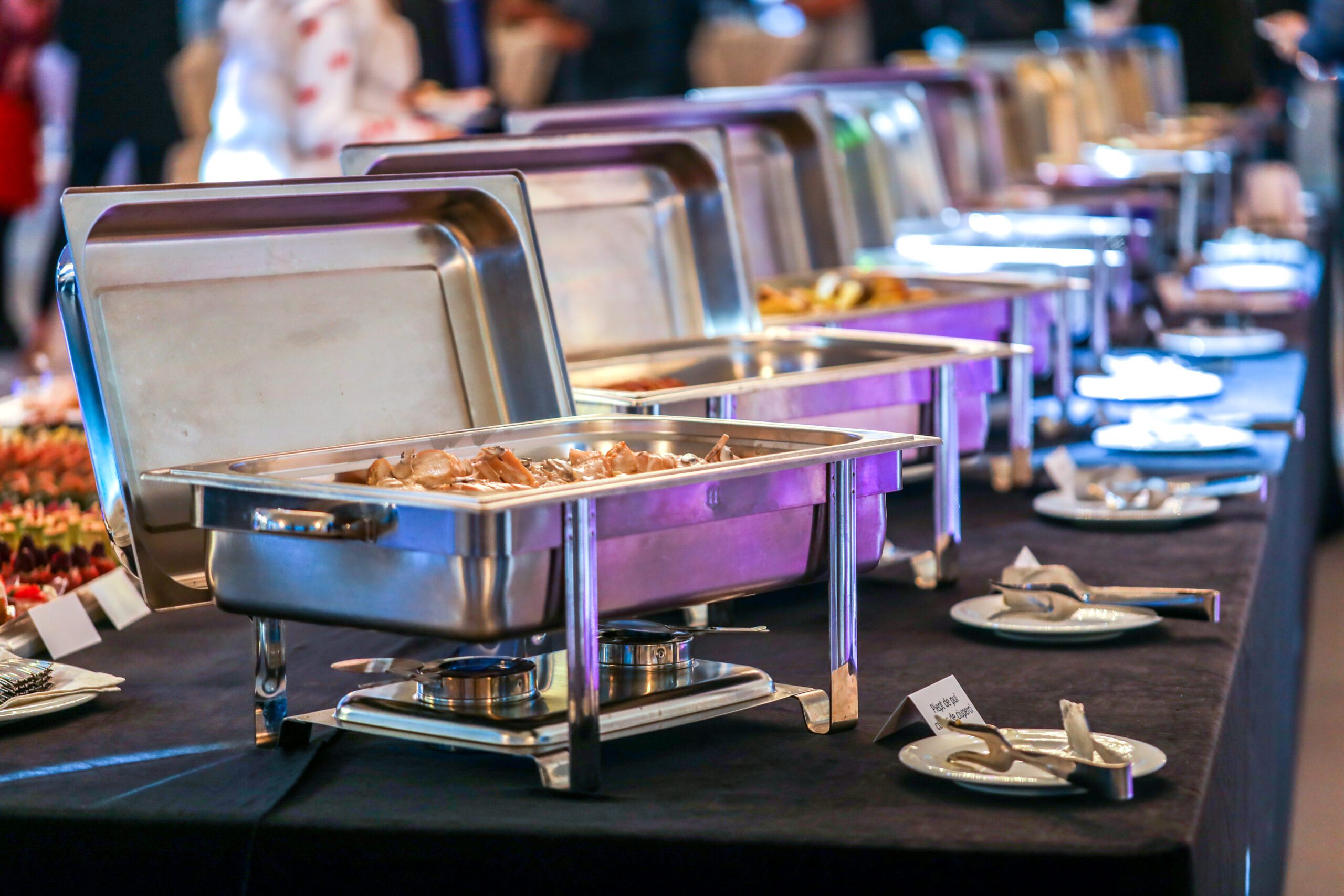 A buffet table with multiple chafing dishes containing various foods, alongside plates and cutlery, set up in a banquet setting.