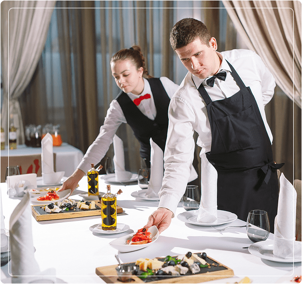 Two servers in uniform set a dining table with white linens, elegant tableware, and food platters.