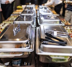 A long table with several closed stainless steel chafing dishes, each with black tongs on top, ready for a buffet. People in the background are standing near the table.