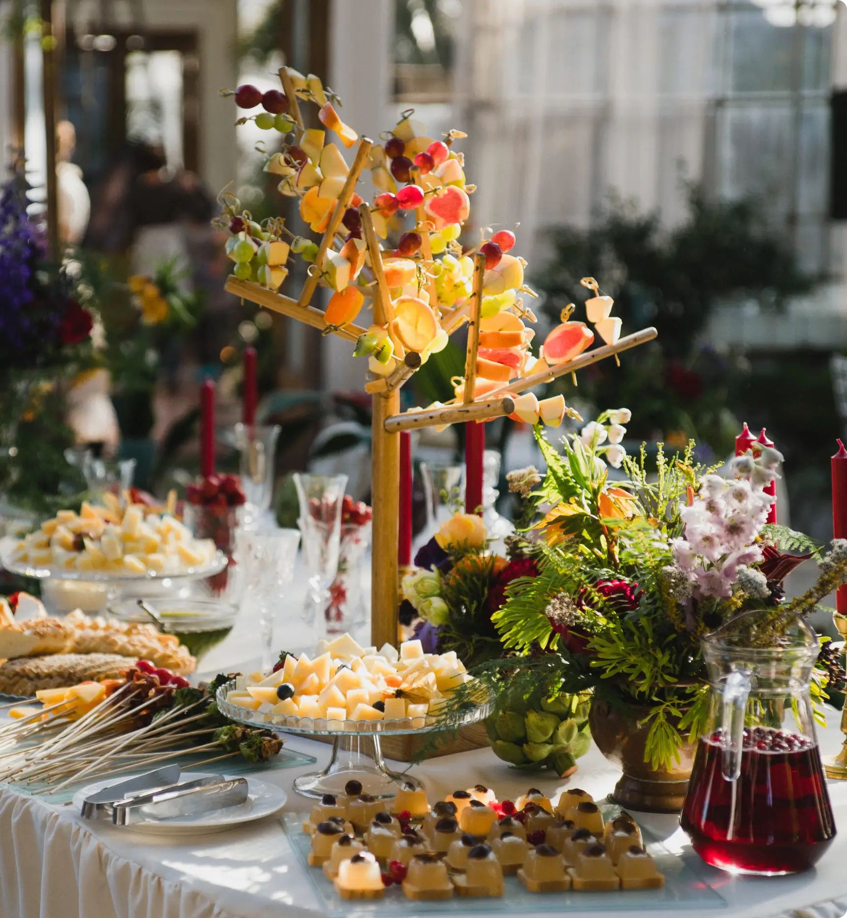 Elegant buffet table with a centerpiece featuring skewered fruits and vegetables. Surrounding dishes include cheese cubes, assorted pastries, and a jug of red beverage.