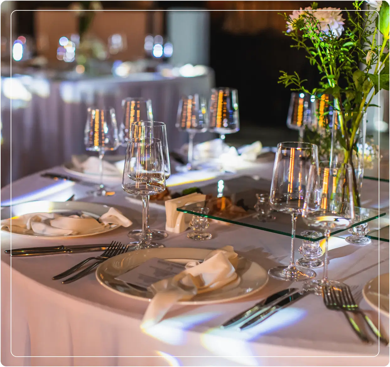 Elegant table set for an event with white tablecloth, folded napkins, glassware, cutlery, and floral centerpieces.