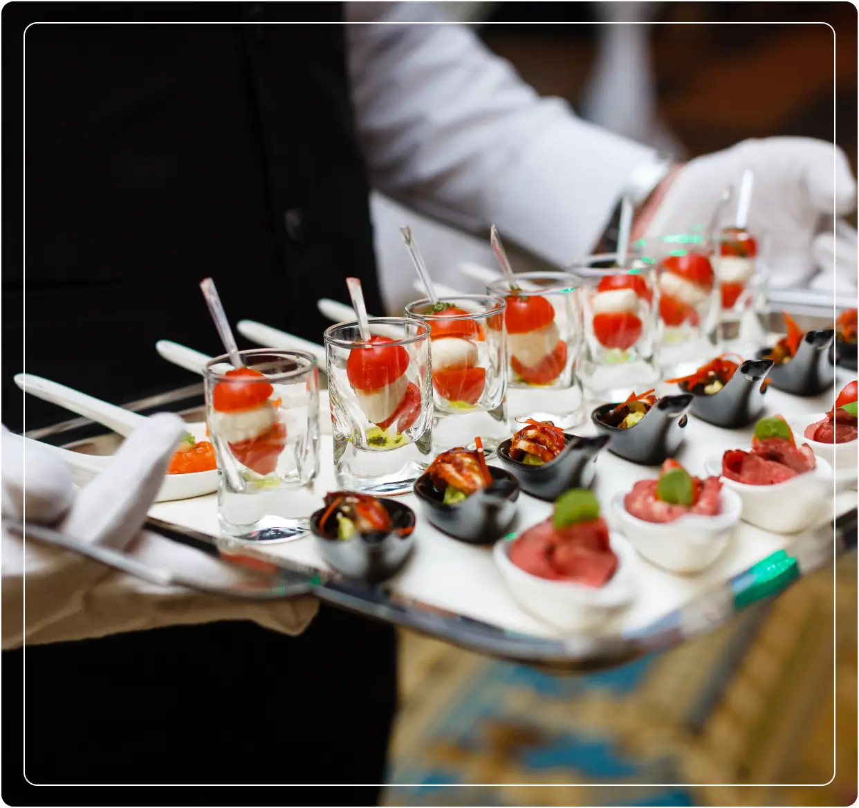 A server in a black vest and white gloves carries a tray with small appetizers, including tomato and mozzarella in glasses and other gourmet bites in small bowls.